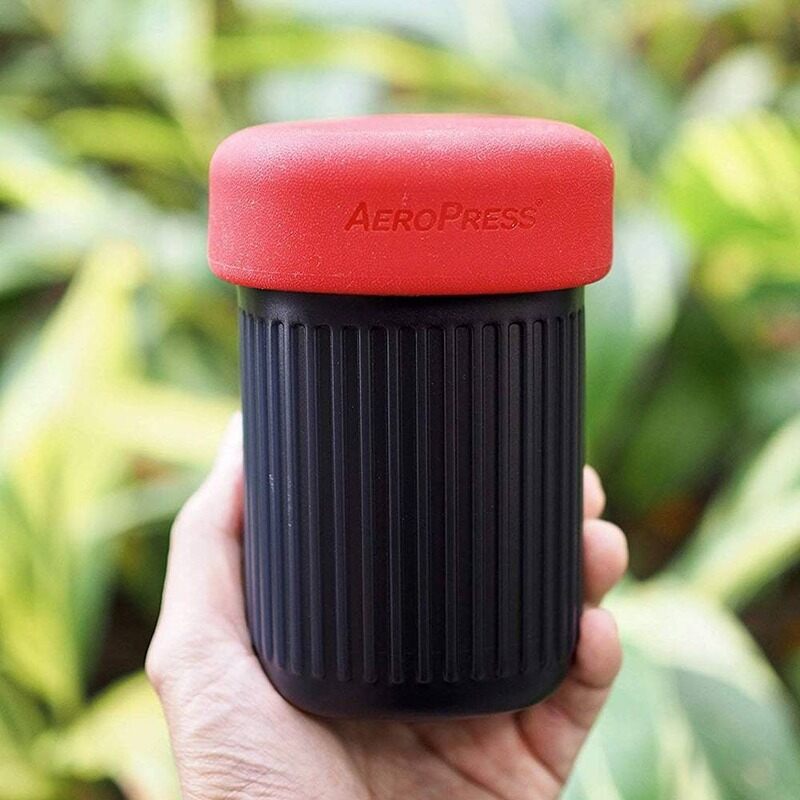 Hand holding the AeroPress Go travel mug with the red silicone lid on, outdoor background blurred.