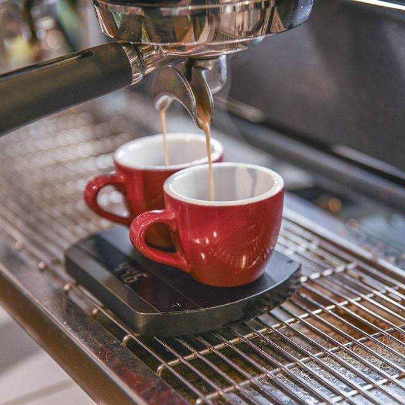 Two red espresso cups under a portafilter, with espresso flowing into each cup, placed on a digital coffee scale