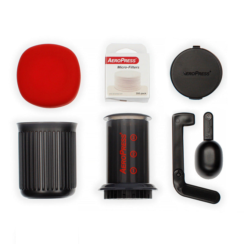 Full AeroPress Go kit laid out, including brewer, red lid, travel mug, scoop, stirrer, filter holder, and pack of micro-filters.