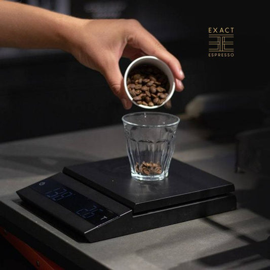 A hand pouring coffee beans into a glass placed on a digital scale.